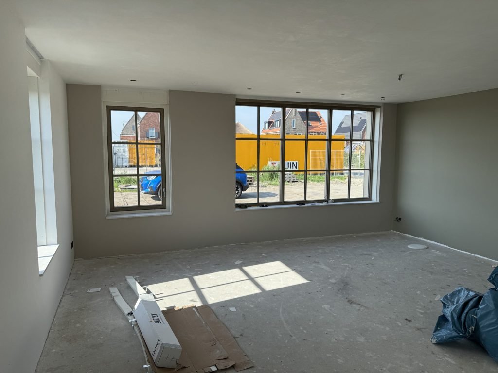 Lege nieuwbouwkamer met wanden in een warme grijstint en grote ramen met zicht op huizen in aanbouw