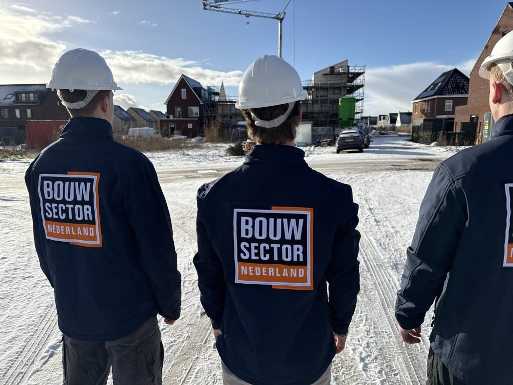 Drie vakmensen van Bouwsector Nederland met witte helm in een besneeuwde nieuwbouwstraat met huizen in aanbouw